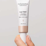 A hand holds Madara H2O Tint Hyaluron Tinted Serum 30ml in shade Moon Flower, a hyaluronic acid-infused tinted moisturiser, against a neutral background.
