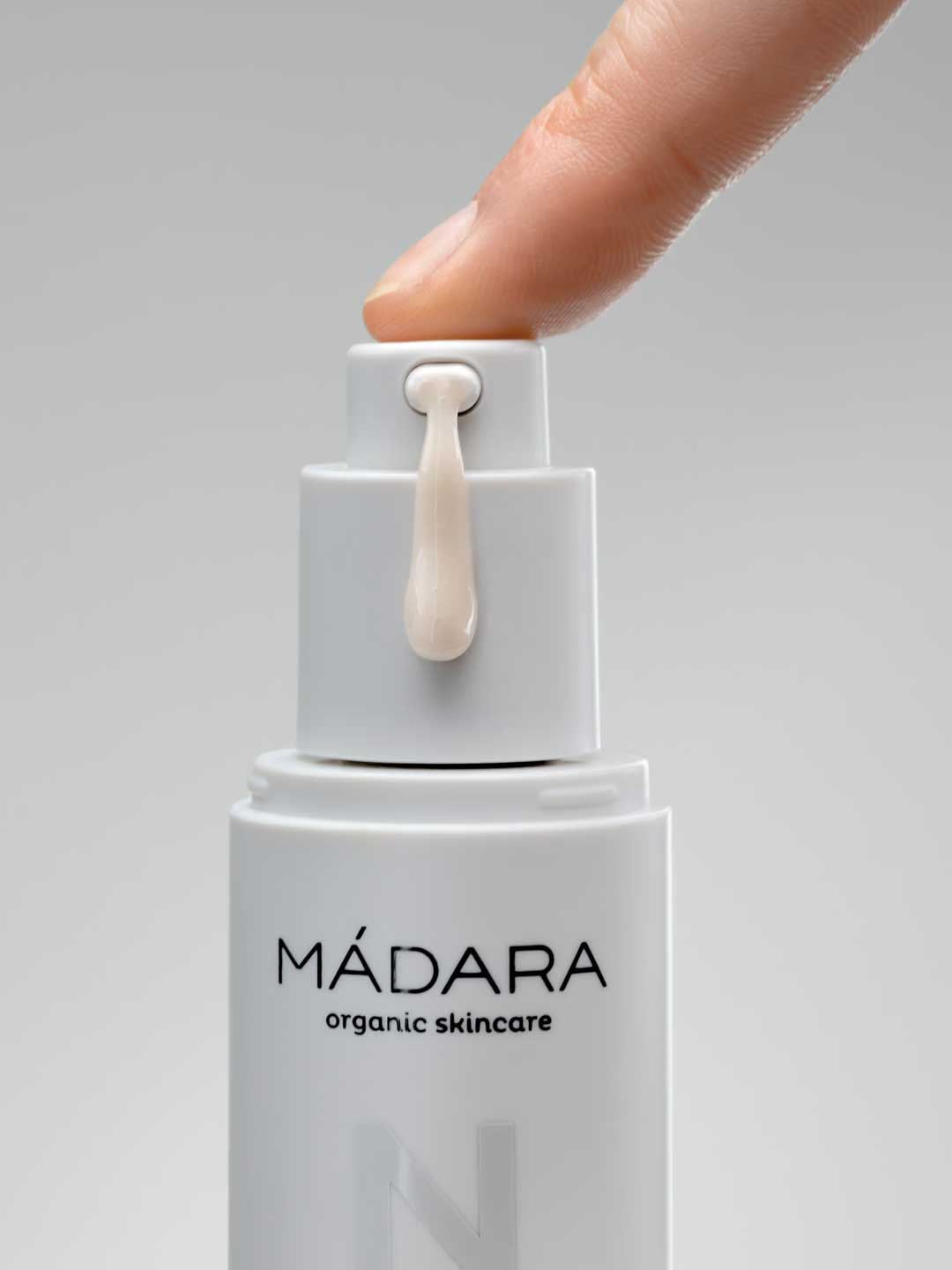 A close-up of a hand pressing the pump of a white Madara Niacinamide Alternative 5-in-1 Serum 30ml bottle, dispensing a drop of serum for breakout-prone skin, set against a plain gray background.