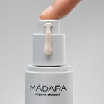 A close-up of a hand pressing the pump of a white Madara Niacinamide Alternative 5-in-1 Serum 30ml bottle, dispensing a drop of serum for breakout-prone skin, set against a plain gray background.