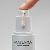 A close-up of a hand pressing the pump of a white Madara Niacinamide Alternative 5-in-1 Serum 30ml bottle, dispensing a drop of serum for breakout-prone skin, set against a plain gray background.
