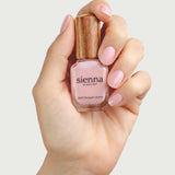 A hand with manicured, light pink nails holds a bottle of Sienna Byron Bay Nail Polish - Magic, featuring a wooden cap, against a plain light background.