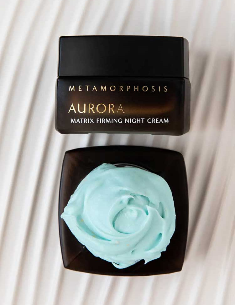 A jar of Metamorphosis Aurora Matrix Firming Night Cream 50ml with its lid off reveals light blue cream inside, resting on a white textured surface.