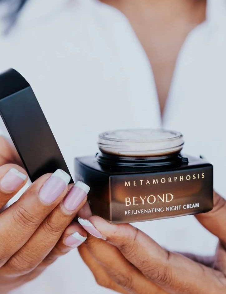 A person with light pink French-manicured nails holds an open jar of Metamorphosis Beyond Rejuvenating Night Cream 50ml, ideal for menopausal skin, with the lid in their other hand and a white shirt in the background.