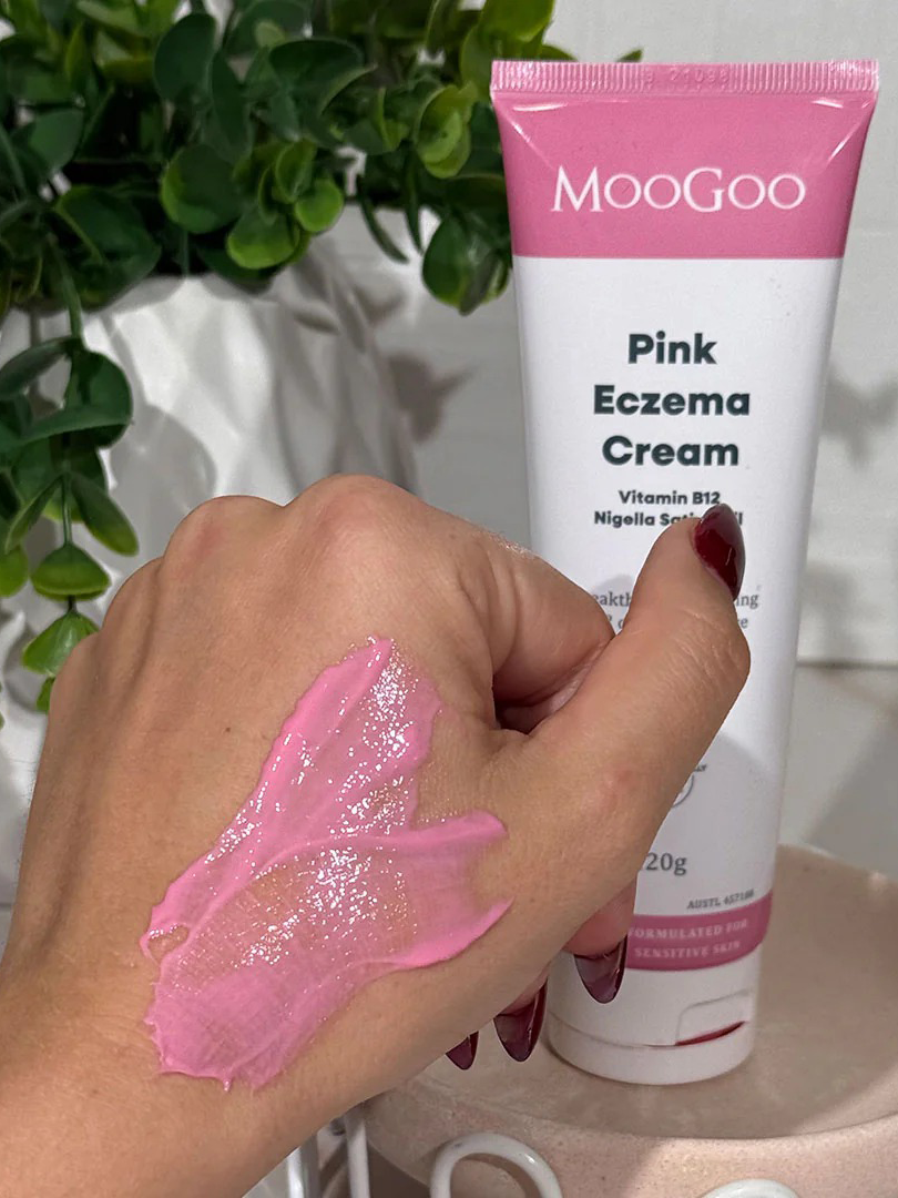 A hand with pink cream is shown in front of a tube labeled MooGoo Pink Eczema Cream 120g by MooGoo, a soothing eczema and dermatitis relief cream. Green plants and various items can be seen in the background.