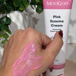 A hand with pink cream is shown in front of a tube labeled MooGoo Pink Eczema Cream 120g by MooGoo, a soothing eczema and dermatitis relief cream. Green plants and various items can be seen in the background.