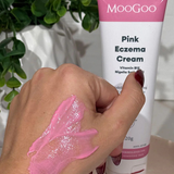 A hand with pink cream is shown in front of a tube labeled MooGoo Pink Eczema Cream 120g by MooGoo, a soothing eczema and dermatitis relief cream. Green plants and various items can be seen in the background.