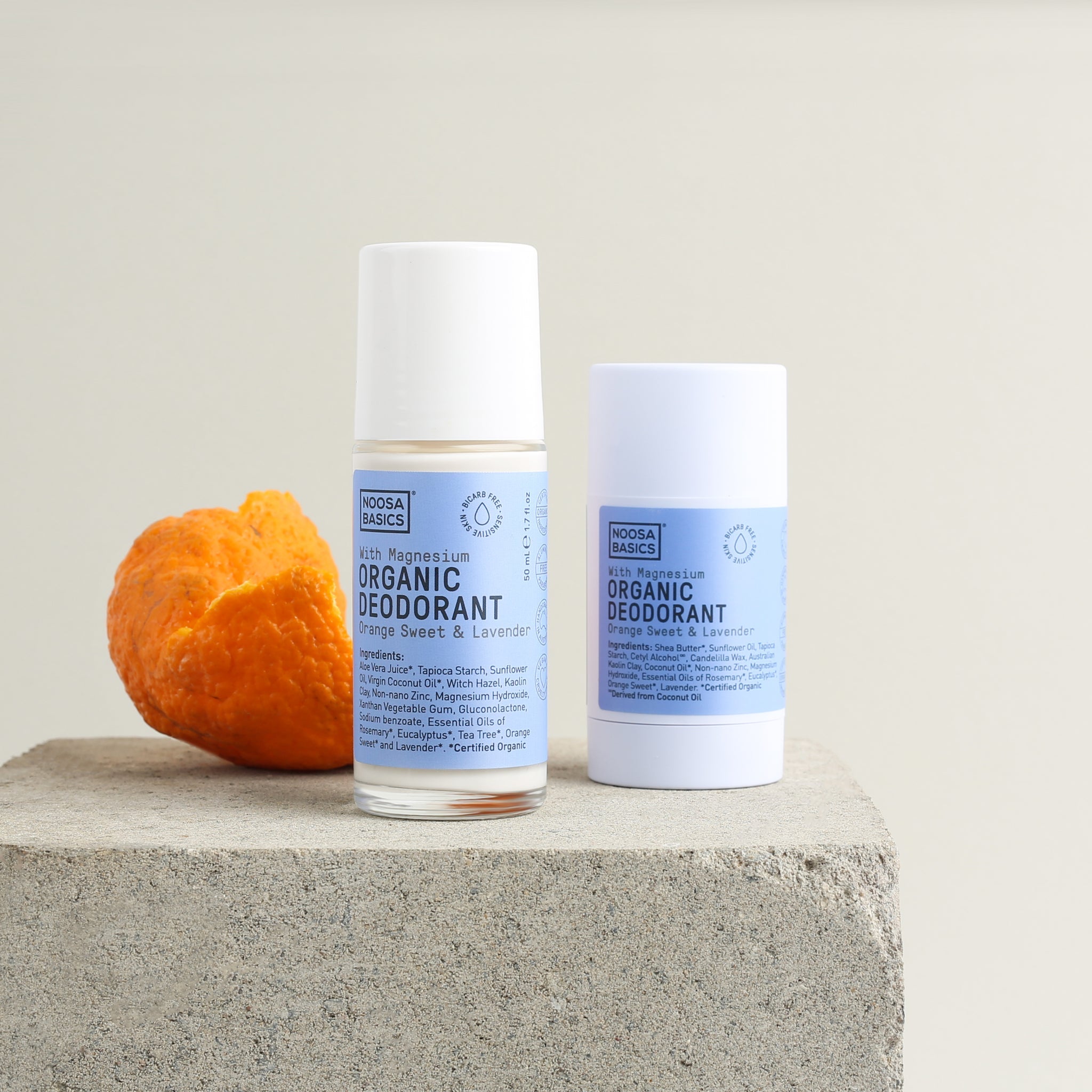 Noosa Basics Sweet Orange and Lavender roll-on and stick deodorants with pale blue labels and white caps, standing on a square stone block beside curled orange peel.