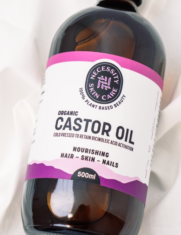 Close-up of an amber Necessity Organic Castor Oil 500ml bottle lying diagonally on white fabric, showing its white label with pink and purple bands.