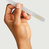 A hand holds the Sienna Byron Bay Glass Nail File, known for its durability and eco-friendly design, with "sienna" printed in yellow on one end against a plain light background.