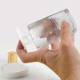 A hand pours clear liquid from a bottle labeled “Sienna Byron Bay Strengthening Nail Polish Remover” onto a cotton pad. Made with plant-derived ingredients, it sits on a white surface with cotton pads and a gold cap nearby.