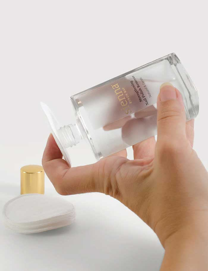 A hand pours clear liquid from a bottle labeled “Sienna Byron Bay Strengthening Nail Polish Remover” onto a cotton pad. Made with plant-derived ingredients, it sits on a white surface with cotton pads and a gold cap nearby.