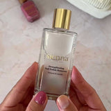 A person holds a clear glass bottle of Sienna Byron Bay Strengthening Nail Polish Remover with a gold cap. Made from plant-derived ingredients, it rests in front of mauve nail polish and a Sienna-branded pouch.