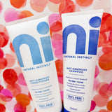 Two white and blue tubes of the Natural Instinct Anti-Dandruff Pack are held against a vibrant pink and red abstract background. Each 250ml tube is labeled as 100% free from harsh chemicals.