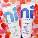 Hand holding two white Natural Instinct Anti-Dandruff tubes against a pink, red and orange spotted background; the conditioner has turquoise lettering and the shampoo has dark blue lettering.