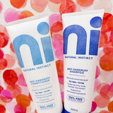 Hand holding two white Natural Instinct Anti-Dandruff tubes against a pink, red and orange spotted background; the conditioner has turquoise lettering and the shampoo has dark blue lettering.