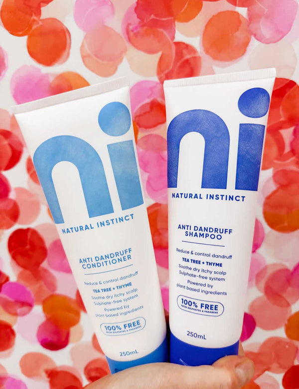 Hand holding two white Natural Instinct Anti-Dandruff tubes against a pink, red and orange spotted background; the conditioner has turquoise lettering and the shampoo has dark blue lettering.