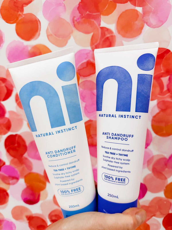 Hand holding two white Natural Instinct Anti-Dandruff tubes against a pink, red and orange spotted background; the conditioner has turquoise lettering and the shampoo has dark blue lettering.