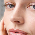 Close-up of a light-skinned face with blue eyes, natural makeup, and soft pink-brown lips. Their hand rests on their cheek. Text reads: Ere Perez Acai Lip Pencils 1.1g in shade Naughty | rose brown, a hydrating liner for added definition.