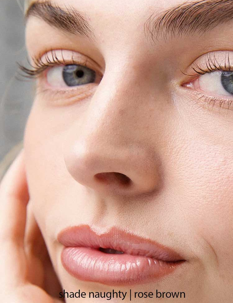 Close-up of a light-skinned face with blue eyes, natural makeup, and soft pink-brown lips. Their hand rests on their cheek. Text reads: Ere Perez Acai Lip Pencils 1.1g in shade Naughty | rose brown, a hydrating liner for added definition.