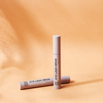 Two slim beige Noosa Basics Eye Lash and Brow Revitaliser tubes against orange, one standing upright and one lying behind it, with long shadows across the surface.