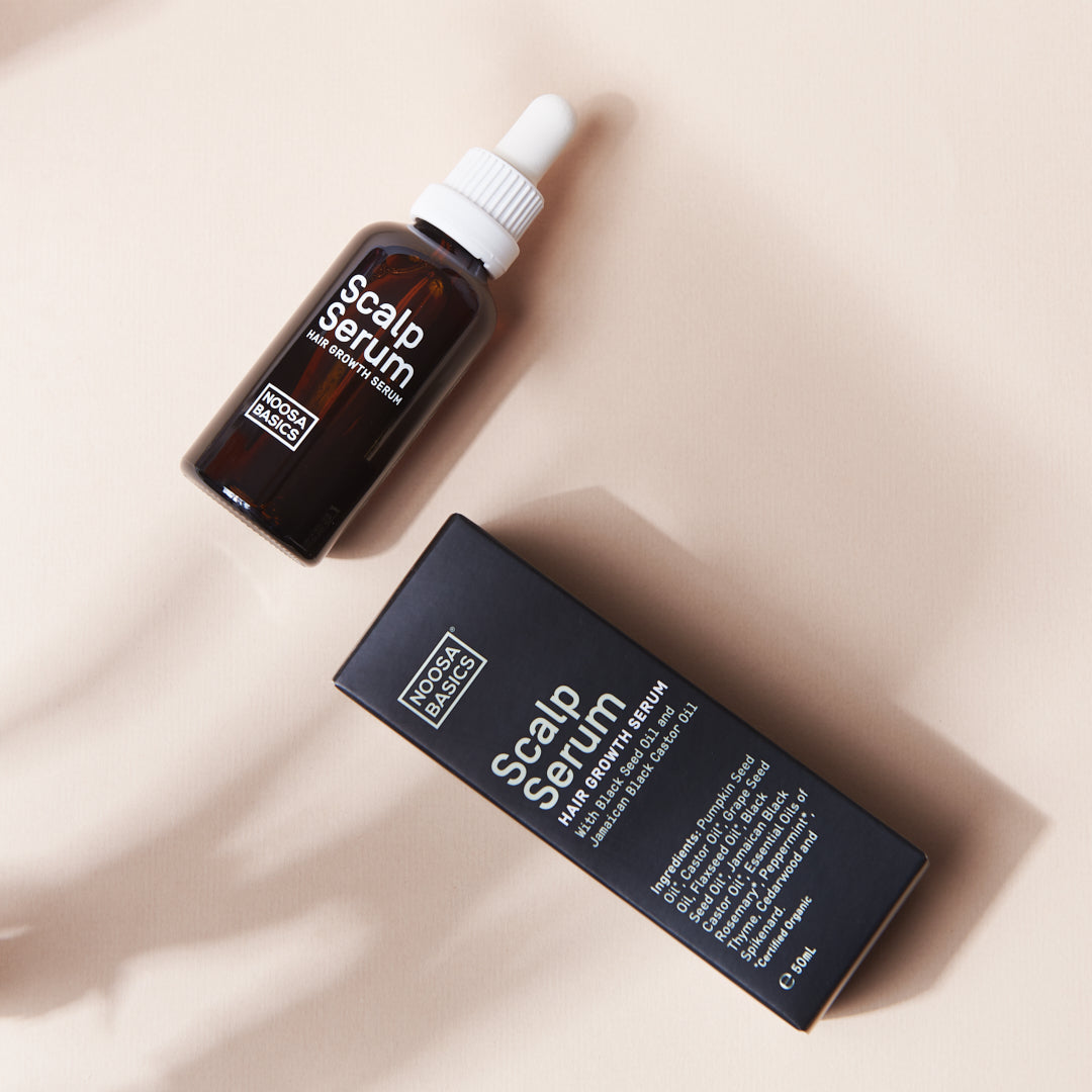 Amber Noosa Basics Scalp Serum bottle with a white dropper cap lying diagonally beside its black carton on a pale surface crossed by soft shadows.