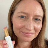 A woman with straight light brown hair smiles while holding a Luk Beautifood Lip Nourish Lipstick 3g. The soft nude shade is made with food-grade ingredients. "Nude Sugar" appears at the bottom against a plain background.