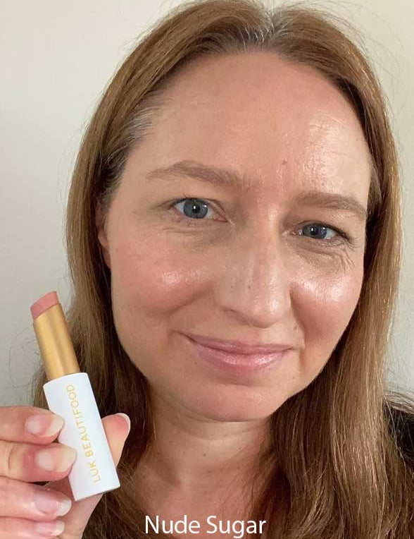 A woman with straight light brown hair smiles while holding a Luk Beautifood Lip Nourish Lipstick 3g. The soft nude shade is made with food-grade ingredients. "Nude Sugar" appears at the bottom against a plain background.