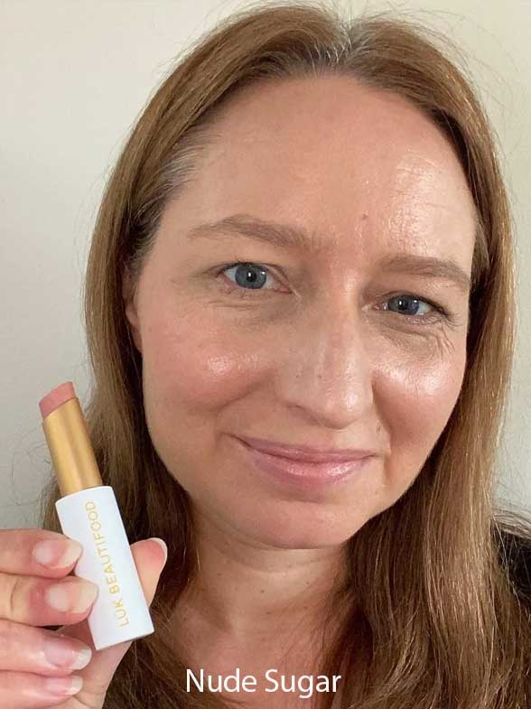 A woman with straight light brown hair smiles while holding a Luk Beautifood Lip Nourish Lipstick 3g. The soft nude shade is made with food-grade ingredients. "Nude Sugar" appears at the bottom against a plain background.
