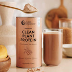 A can of Nutra Naturals Clean Plant Protein Creamy Chocolate 500g sits beside a glass of chocolate shake. A spoonful of protein powder floats above, with chickpeas and fruit scattered in the background.