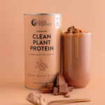 A container of Nutra Naturals Clean Plant Protein Creamy Chocolate 500g stands next to a glass of chocolate protein shake topped with chocolate pieces, with chocolate chunks and a spoonful of protein powder in front.