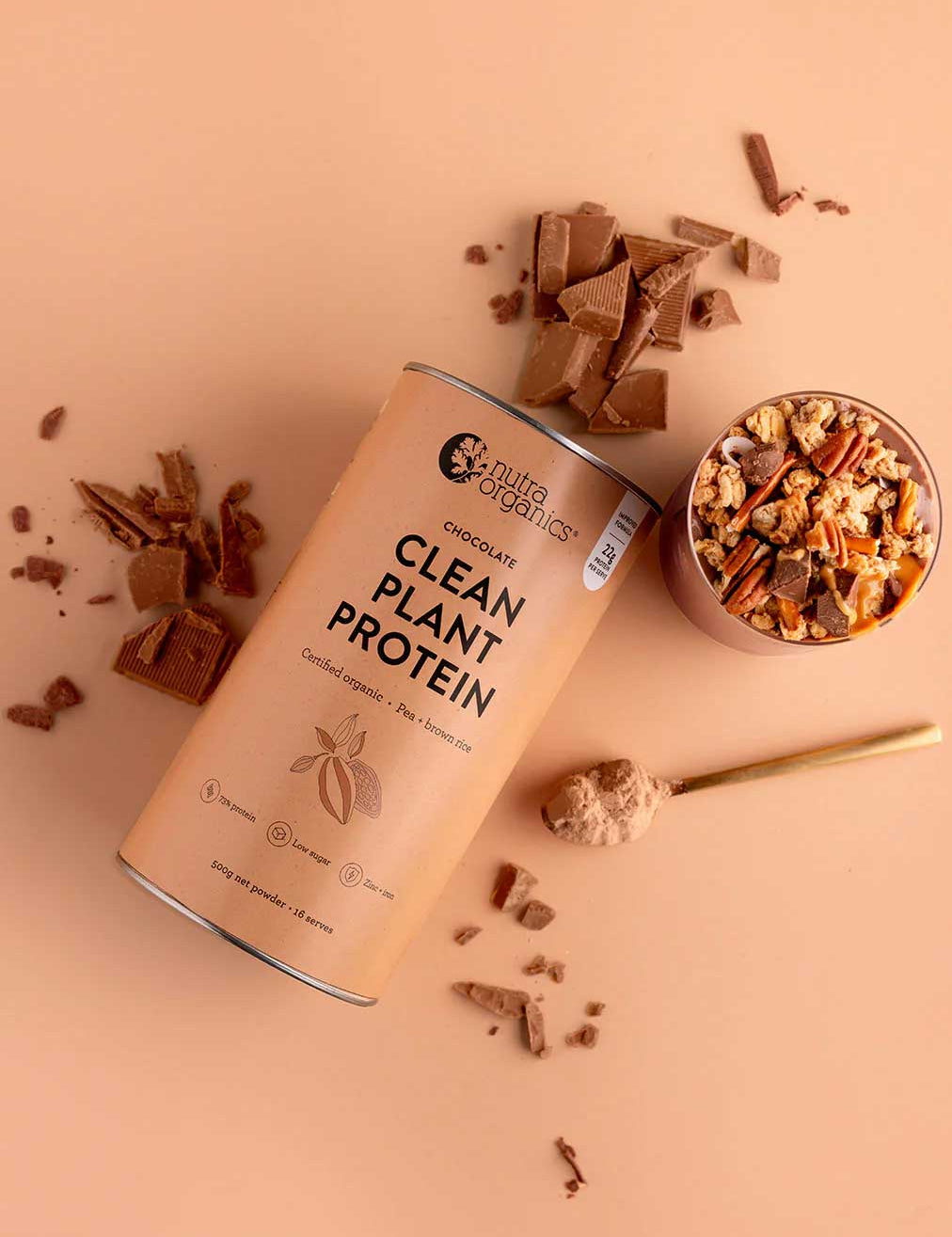 A canister of Nutra Naturals Clean Plant Protein Creamy Chocolate 500g sits on a beige surface beside broken chocolate pieces, a bowl of granola, and a gold spoon with vegan protein powder.