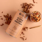 A canister of Nutra Naturals Clean Plant Protein Creamy Chocolate 500g sits on a beige surface beside broken chocolate pieces, a bowl of granola, and a gold spoon with vegan protein powder.