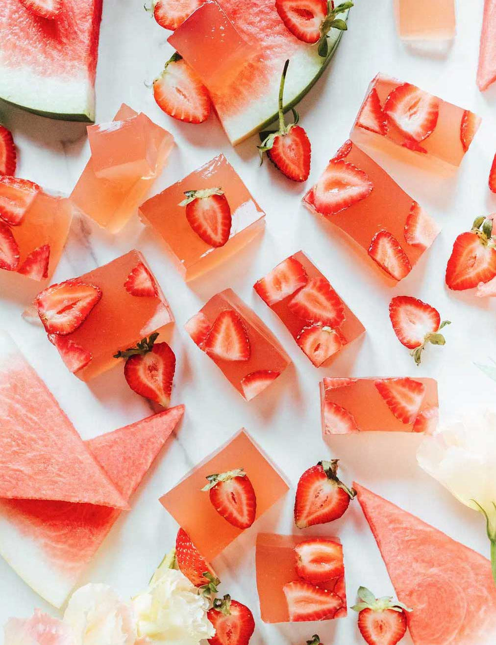 Cubed pink Nutra Naturals Collagen Beauty Watermelon Strawberry desserts with sliced strawberries rest on a white surface, surrounded by fresh watermelon triangles, a bright, refreshing setup perfect for a nourishing treat.