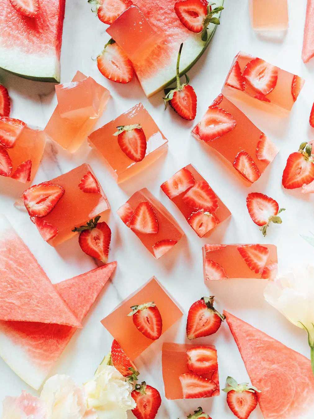 Cubed pink Nutra Naturals Collagen Beauty Watermelon Strawberry desserts with sliced strawberries rest on a white surface, surrounded by fresh watermelon triangles—a bright, refreshing setup perfect for a nourishing treat.