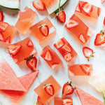 Cubed pink Nutra Naturals Collagen Beauty Watermelon Strawberry desserts with sliced strawberries rest on a white surface, surrounded by fresh watermelon triangles—a bright, refreshing setup perfect for a nourishing treat.