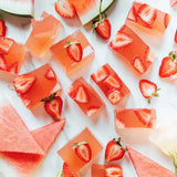 Cubed pink Nutra Naturals Collagen Beauty Watermelon Strawberry desserts with sliced strawberries rest on a white surface, surrounded by fresh watermelon triangles—a bright, refreshing setup perfect for a nourishing treat.