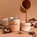 A hand pours creamy Nutra Naturals Collagen Hot Chocolate 200g into a mug next to its tin, with chocolate chunks and cocoa powder scattered around, creating a cozy scene for relaxation on a beige background.