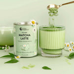 A glass of Nutra Naturals Collagen Matcha Latte 100g is paired with its can, as a spoon sprinkles organic Japanese matcha over green leaves and white flowers on a pale green background.