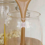Nutra Naturals Collagen Protein Unflavoured, a creamy light brown liquid, is poured into a clear glass jar with rolled oats in the background—ideal for muscle recovery thanks to added collagen protein.