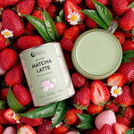 A tin of Nutra Naturals Collagen Strawberry Matcha Latte 100g, a mug of latte, strawberries, green leaves, and white flowers are arranged on a bed of fresh strawberries.