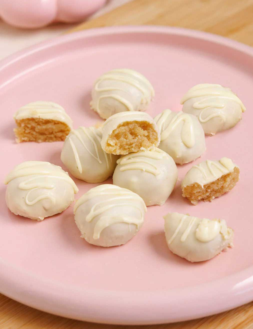 Nutra Naturals Collagen White Hot Choc Salted Caramel 200g cake balls are coated in white chocolate and low in sugar, with a golden, moist interior. Some are whole, some cut open, and all are arranged on a pink plate atop a wooden surface.