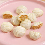 Nutra Naturals Collagen White Hot Choc Salted Caramel 200g cake balls are coated in white chocolate and low in sugar, with a golden, moist interior. Some are whole, some cut open, and all are arranged on a pink plate atop a wooden surface.