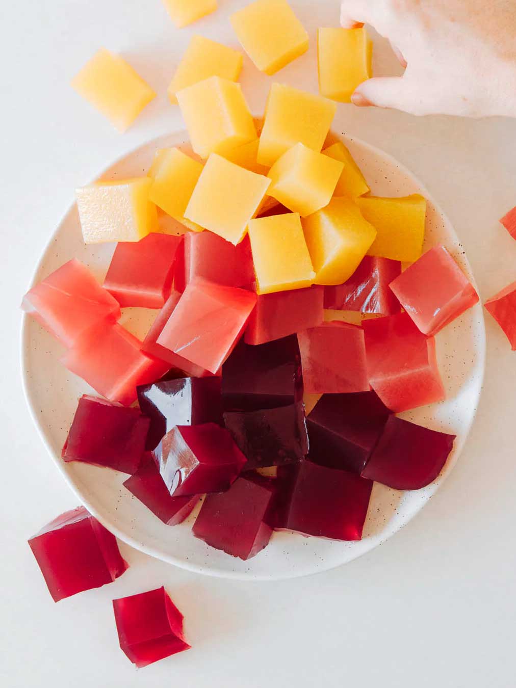 A plate of colorful gelatin cubes in yellow, pink, and dark red on a white surface, with a hand reaching for a yellow cube made using Nutra Naturals Collagen Beauty Blueberry Wildflower 225g for a beauty-boosting treat.