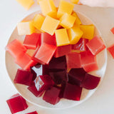 A plate of colorful gelatin cubes in yellow, pink, and dark red on a white surface, with a hand reaching for a yellow cube made using Nutra Naturals Collagen Beauty Blueberry Wildflower 225g for a beauty-boosting treat.