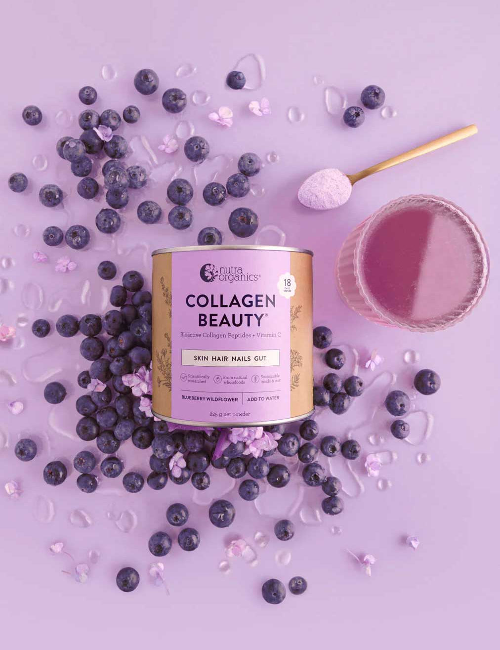 A canister of Nutra Naturals Collagen Beauty Blueberry Wildflower 225g is surrounded by blueberries, purple powder, and a glass of purple drink on a light purple background. A scoop of hydrolysed bovine collagen powder rests nearby.