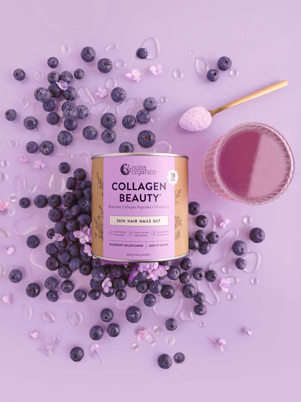 A canister of Nutra Naturals Collagen Beauty Blueberry Wildflower 225g is surrounded by blueberries, purple powder, and a glass of purple drink on a light purple background. A scoop of hydrolysed bovine collagen powder rests nearby.
