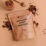 A bag of Nutra Naturals Organic Whey Protein Chocolate 750g sits on a tan surface, surrounded by chocolate pieces, a spoonful of powder, and a glass filled with a rich chocolate protein shake.