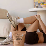A person stretches their legs on an exercise mat with a bag of Nutra Naturals Organic Whey Protein Chocolate 750g near their feet; a sofa and a table with a decorative painting are visible in the background.