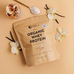 A beige pouch of Nutra Naturals Organic Whey Protein Vanilla 750g, made from grass-fed New Zealand dairy, rests on a light tan surface with white orchids, vanilla beans, a spoon of protein powder, granola, yogurt, and fruit.
