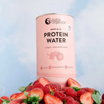A pink Nutra Naturals Protein Water Berry Bliss 450g container sits on fresh strawberries against a soft sky-blue backdrop with clouds, emphasizing its natural electrolytes and refreshing protein water blend.